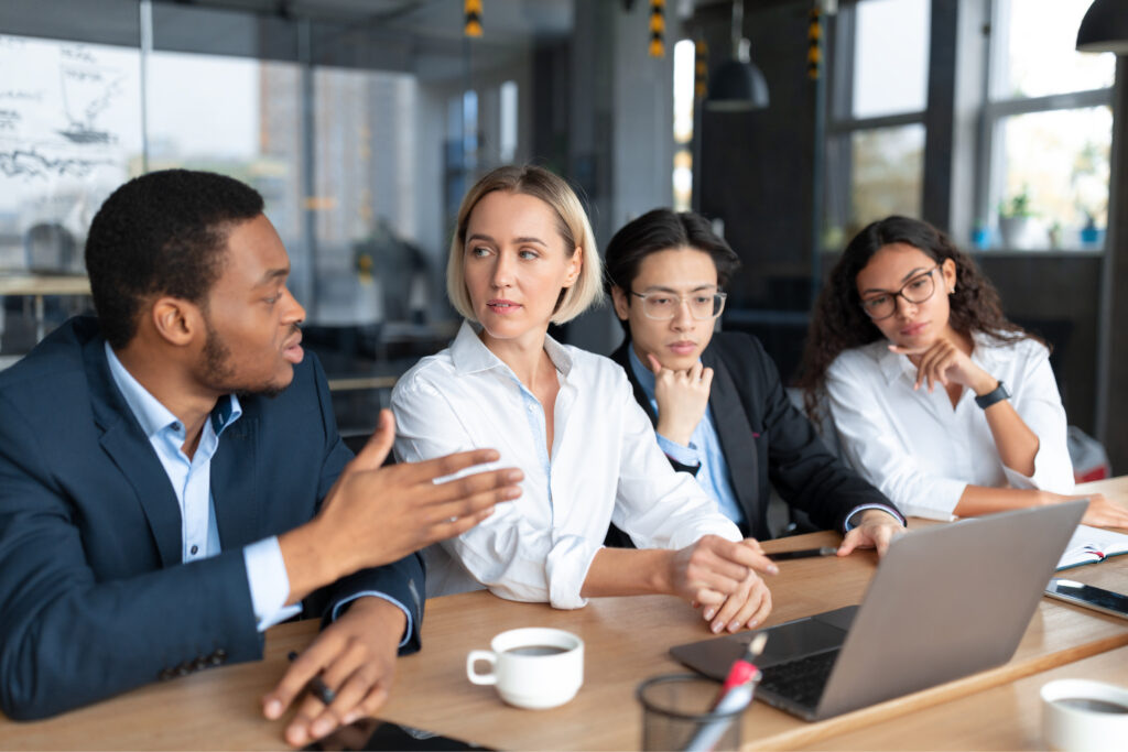 diverse group sits on one side of a table discussing materials on laptop